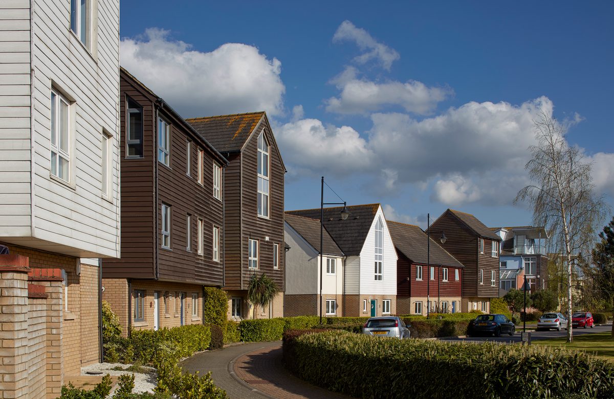 Some of the different coloured, wooden clad houses with gabled rooves of varying heights, set around a landscaped, green public space within the Fishing Village part of St Mary's Island masterplan.