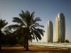 A wide exterior shot of the Al Bahr Towers in Abu Dhabi with the surrounding context in shot, including palm trees in the foreground.
