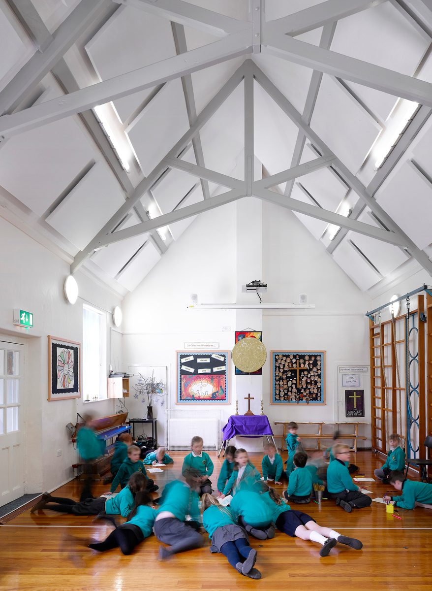 Pupils using the mezzanine floor of the refurbished hall space of All Saints CE Primary School with original beams in the eaves space, white walls and wooden flooring.