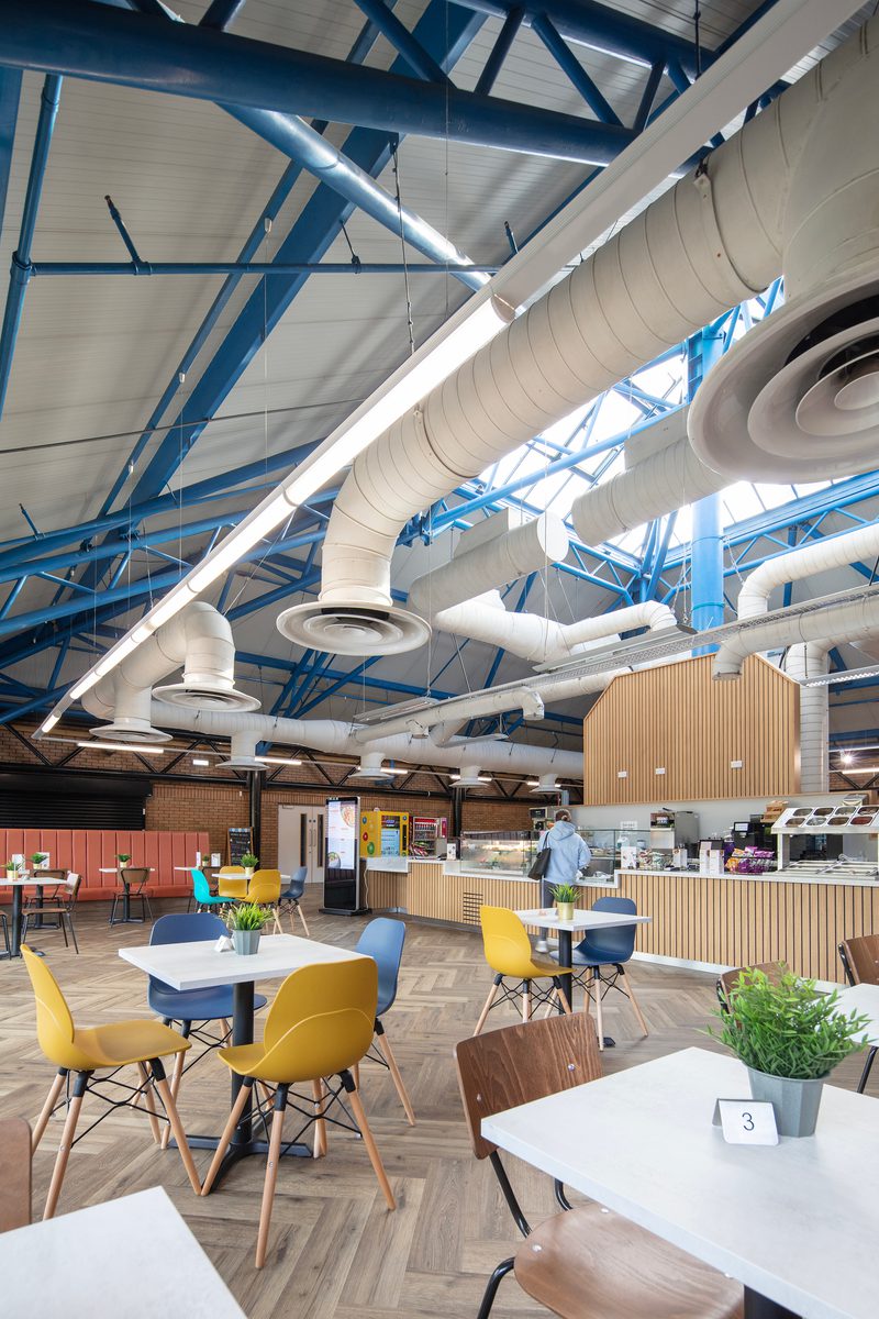 Tables and coloured chairs within the bright reception area of the refurbished Barnsley Metrodome, with café facilities, wooden detailing, roof lights and exposed brickwork.