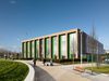 The exterior of the sixth form college within Beswick Community Hub masterplan, showing the building in its entirety, its highly glazed façade and people walking through the landscaped surroundings.