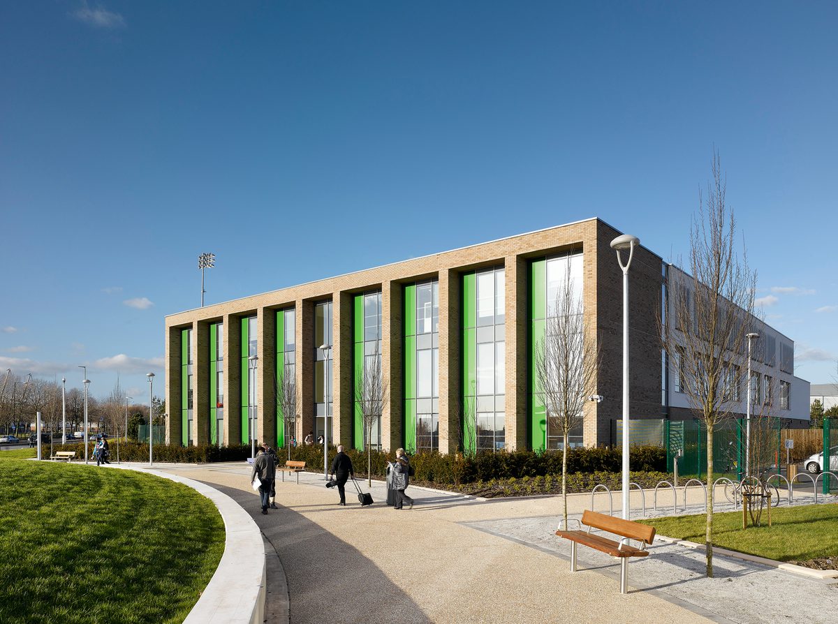 The exterior of the sixth form college within Beswick Community Hub masterplan, showing the building in its entirety, its highly glazed façade and people walking through the landscaped surroundings.