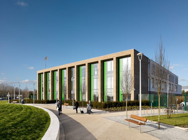 The exterior of the sixth form college within Beswick Community Hub masterplan, showing the building in its entirety, its highly glazed façade and people walking through the landscaped surroundings.