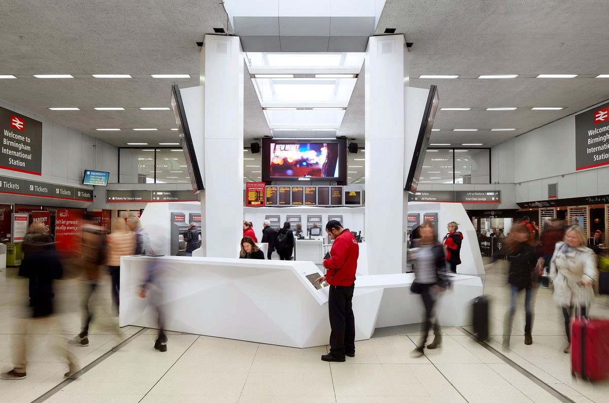 The redeveloped ticket hall at Birmingham International Train Station, with large, white hub of ticket machines in the centre and information boards, other ticket machines and pedestrians surrounding.