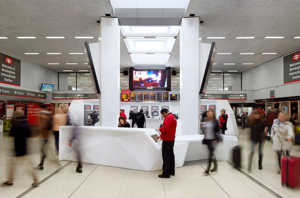 The redeveloped ticket hall at Birmingham International Train Station, with large, white hub of ticket machines in the centre and information boards, other ticket machines and pedestrians surrounding.
