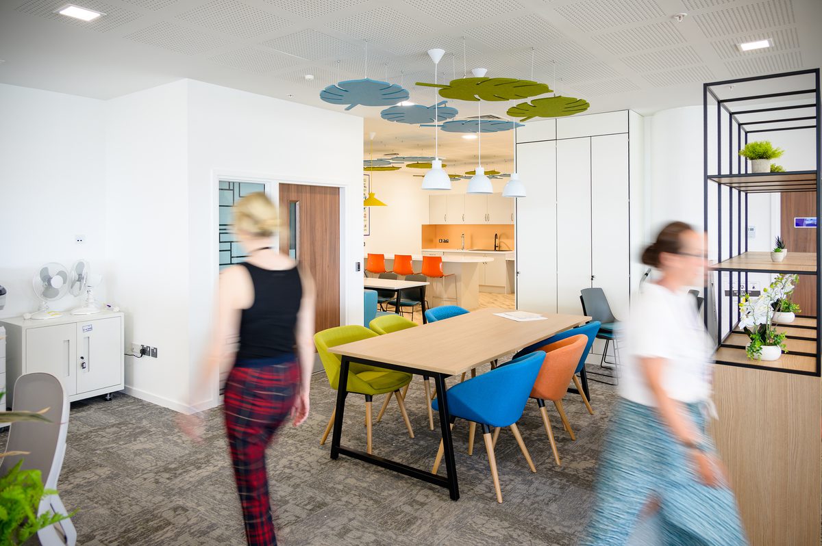 Two people walking through the bright interior of the Children and Family Court Advisory and Support Service office, with kitchen facilities, communal seating and colourful furniture and fixtures.