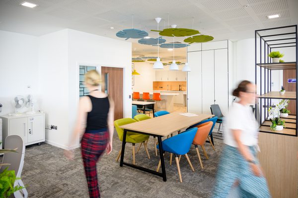 Two people walking through the bright interior of the Children and Family Court Advisory and Support Service office, with kitchen facilities, communal seating and colourful furniture and fixtures.