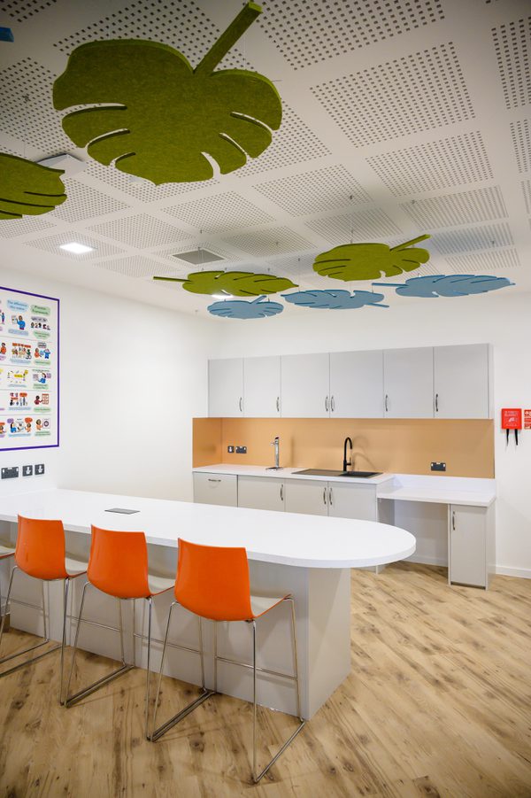 The bright, communal kitchen area within the Children and Family Court Advisory and Support Service office, with colourful furniture and decorative green leaves on the ceiling.