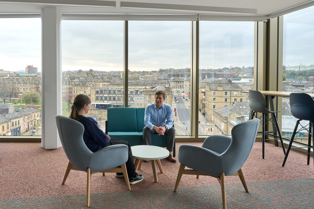 Two colleagues at the university sat down chatting on comfy chairs inside the staff area overlooking Huddersfield.