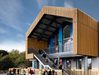 Students walking around the front exterior of Dartmouth Academy, with gabled roof, wooden cladding, blue facade detailing and playground in front.