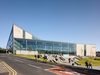 The exterior of Darwen Aldridge Community Academy, with highly glazed facade, distinct triangular roof, landscaped surroundings and students walking in front of the college.