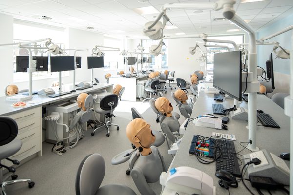 A row of dental phantom head simulators in the Emily Siddon Building, mounted on chairs and workbenches that mimic a real dental operatory. On the desks is computer equipment for students.