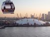One cable car of the Emirates Air Line Cable Car hung from a cable in the top left corner, with a view of the Thames, the O2 Arena and London city centre in the background.