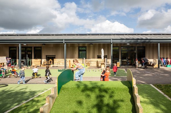 The exterior of Halbeath Nursery, with children playing in green, play space with the wooden clad nursery in the background.