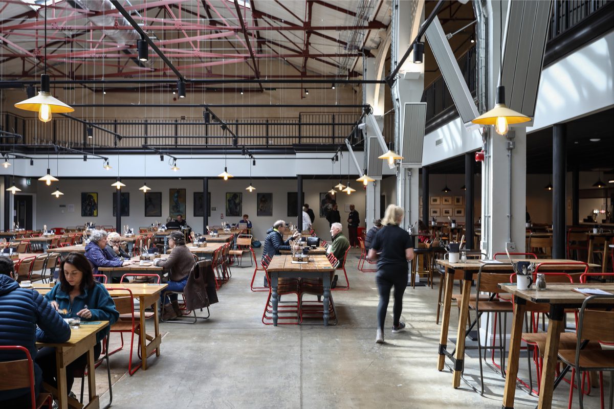 A wide shot of the busy dining space with servers moving across the space and visitors enjoying relaxing across a selection of tables and benches.