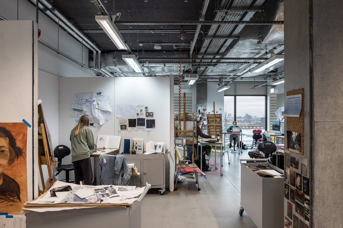 East Quad Building art studio, with simple, robust finishes of plywood walls and vinyl flooring, students are working throughout the space.