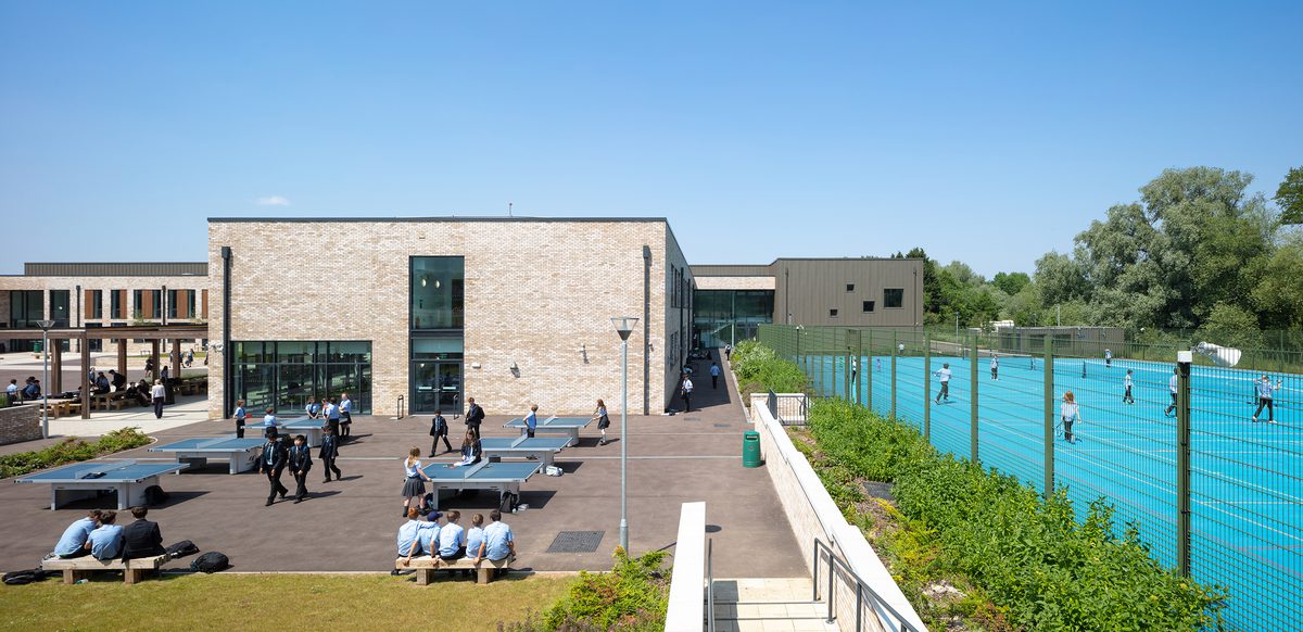 An exterior view of the High School Leckhampton, with hard landscaped area, planting, students socialising and playing games on ping pong tables and students playing on tennis courts to the right.