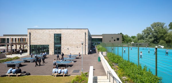 An exterior view of the High School Leckhampton, with hard landscaped area, planting, students socialising and playing games on ping pong tables and students playing on tennis courts to the right.