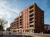 The exterior of the Highgate Road apartment blocks, with red brick façade, large balconies and people moving through the street below.