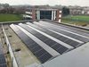 Rows of photovoltaic panels on the roof of Holy Trinity Academy, with school buildings, associated playing fields and suburban surroundings in the background.