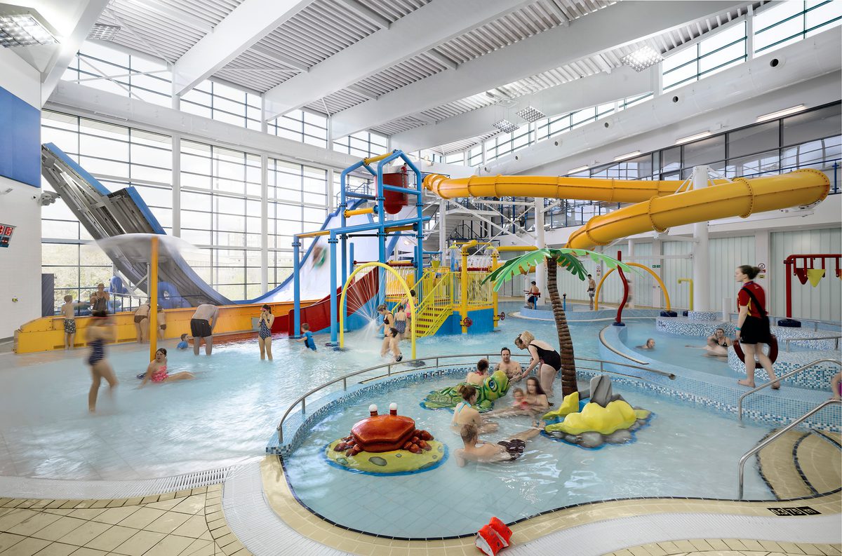 Children playing in the pool hall of Huddersfield Leisure Centre, with highly glazed walls and water slides.