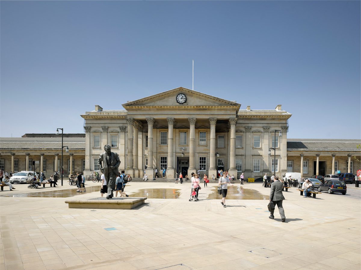The ornate and decorative front entrance of the Grade I listed Huddersfield Train Station with stone columns and clock and pedestrians moving through St George's Square in front.