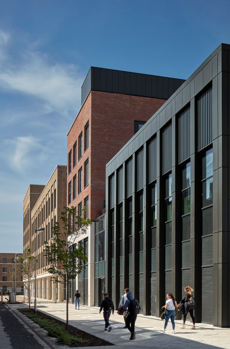 The modern IMPACT building, with brick and dark metal façade along a sunny walkway, with people walking past.