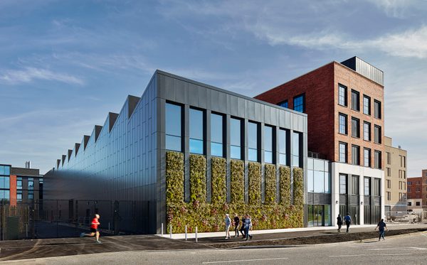 The modern IMPACT building with dark cladding, vertical windows, and a green wall facade with people walking outside.