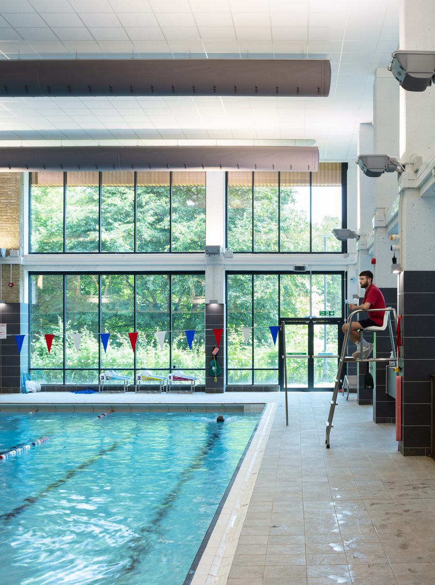 The pool hall within Keynsham Riverside Leisure Centre with highly glazed double height exterior wall, pool and lifeguard watching the swimmers.
