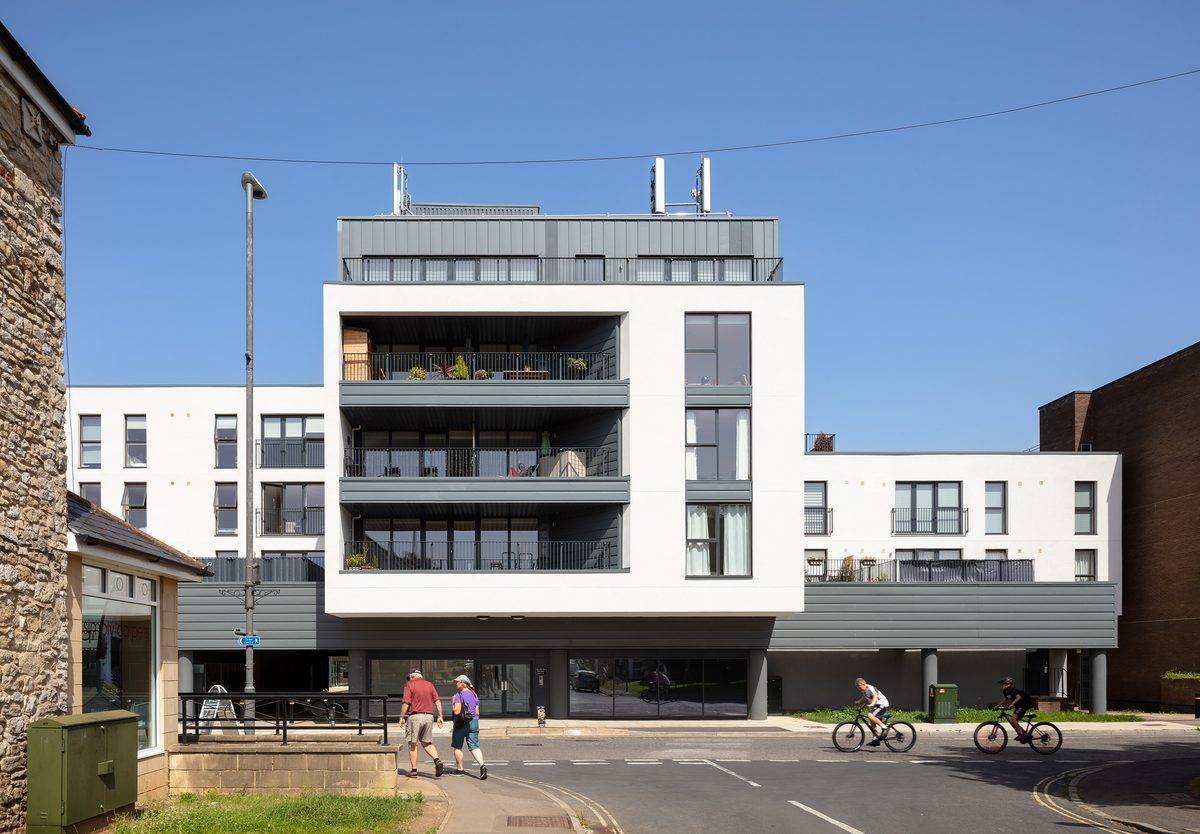 The white exterior of Keynsham Riverside View apartments with balconies and pedestrians walking a street level in front.
