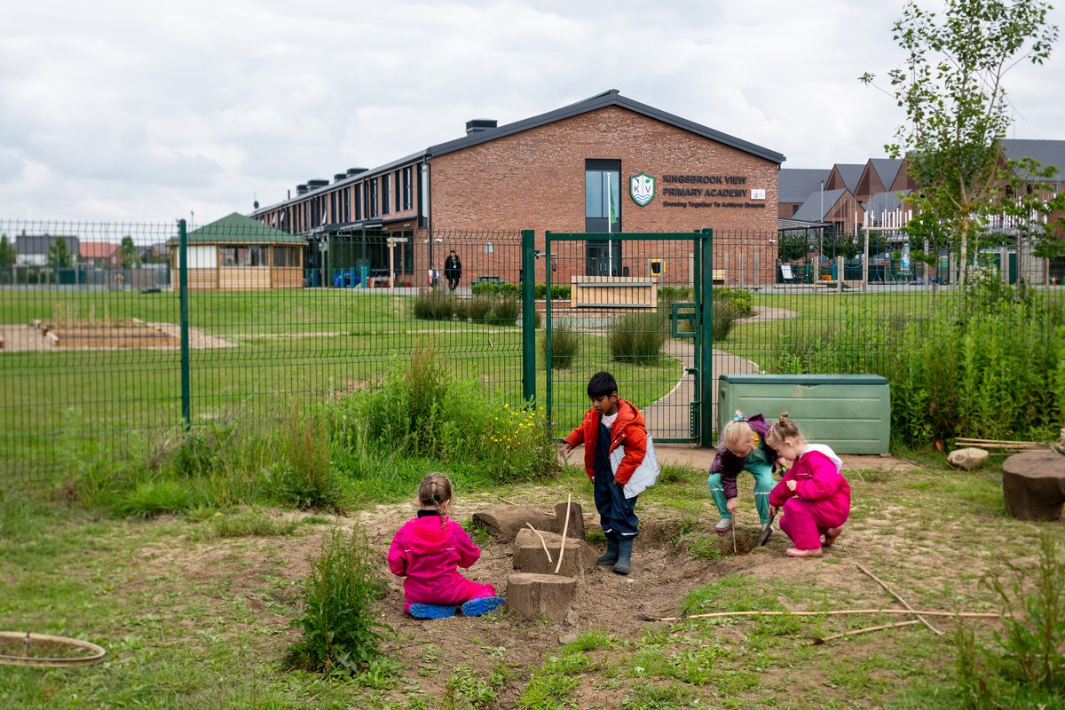 Children playing in the forest school area of Kingsbrook View Primary Academy, surrounded by greenery, planting and secure green fencing, with the school in the background.