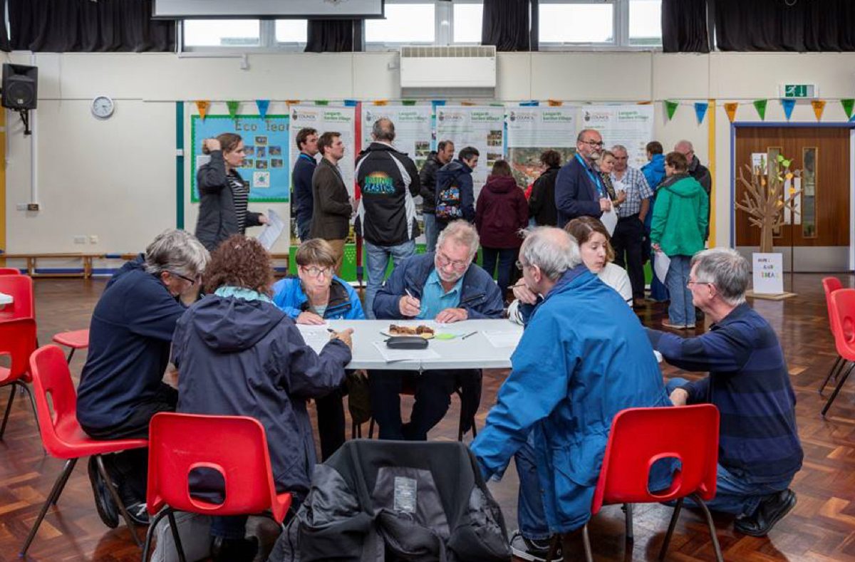 People sat around a table, with more people and visual boards in the background, part of the stakeholder engagement process. used when planning Langarth Garden Village.