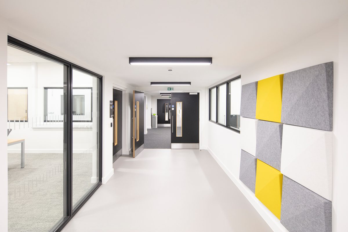 A bright circulation space within the Laura Annie Wilson building, with floor to ceiling glazed, internal windows and decorative yellow, grey and white wall features.