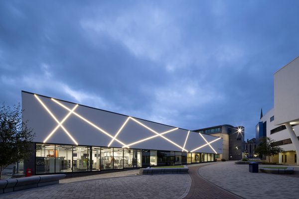 The exterior of the Laura Annie Wilson Building at dusk, with highly glazed ground floor, decorative exterior lightning, surrounded by landscaped public realm.
