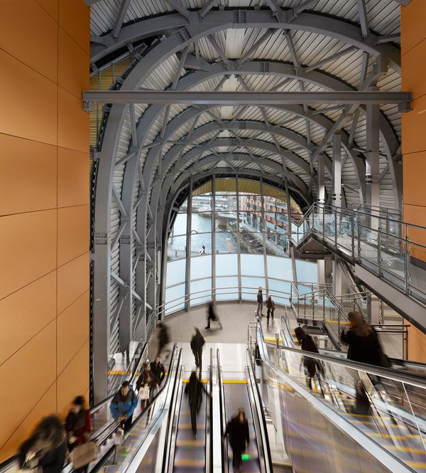 The interior of the Leeds Train Station's southern entrance, with escalators leading down to the entrance's glass front and curved metal beams above.