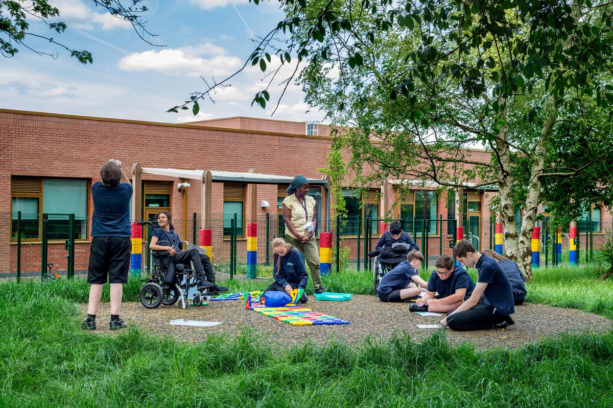 A wide external shot of pupils and a teacher engaging and learning in the outdoor space surrounding by trees, nature and greenery.