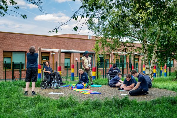 A wide external shot of pupils and a teacher engaging and learning in the outdoor space surrounding by trees, nature and greenery.
