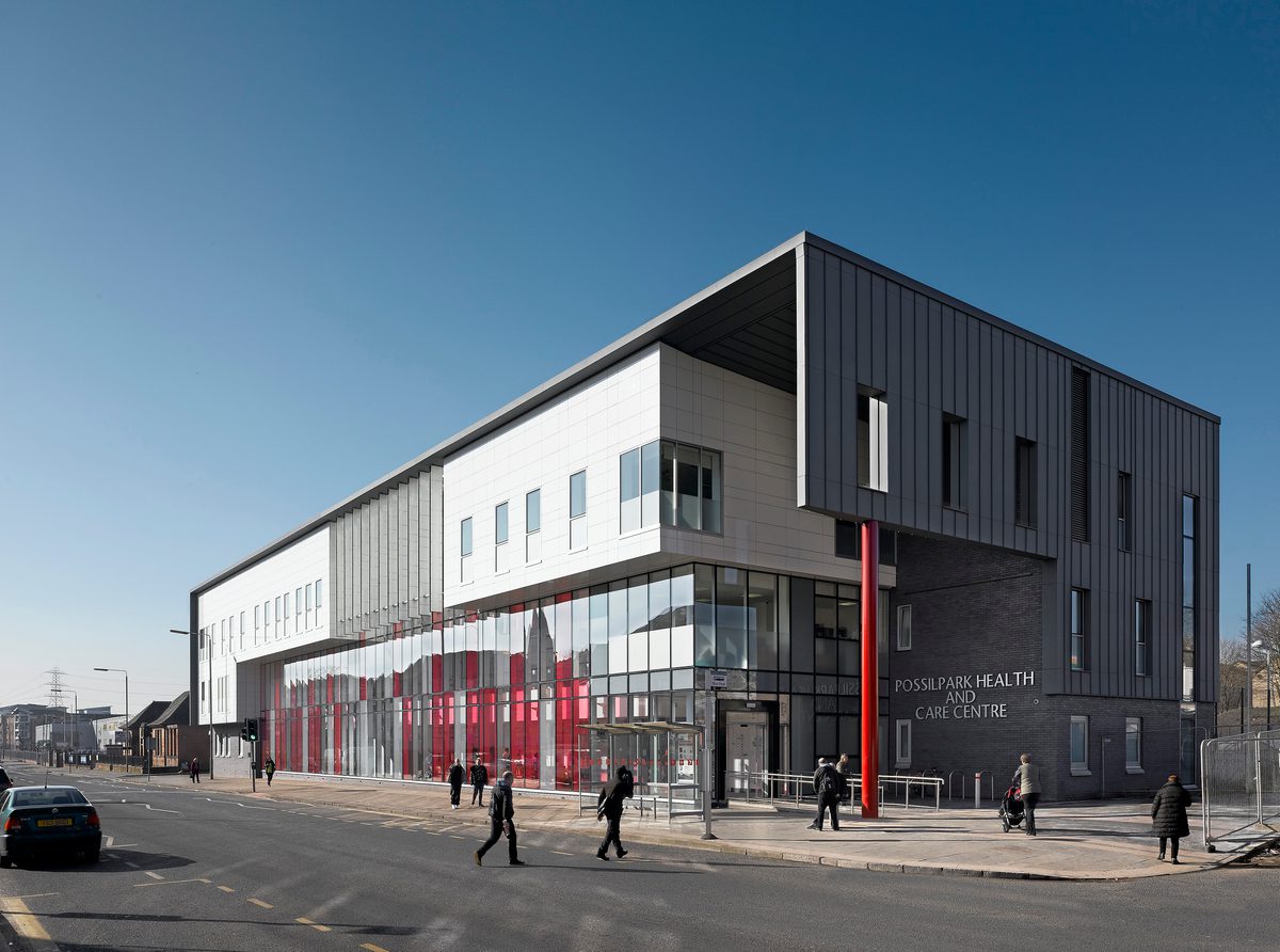 The completed exterior of Possilpark Health Centre in the daytime. With glass cladding and full height ruby red glazing and blue brick running the length of the building.