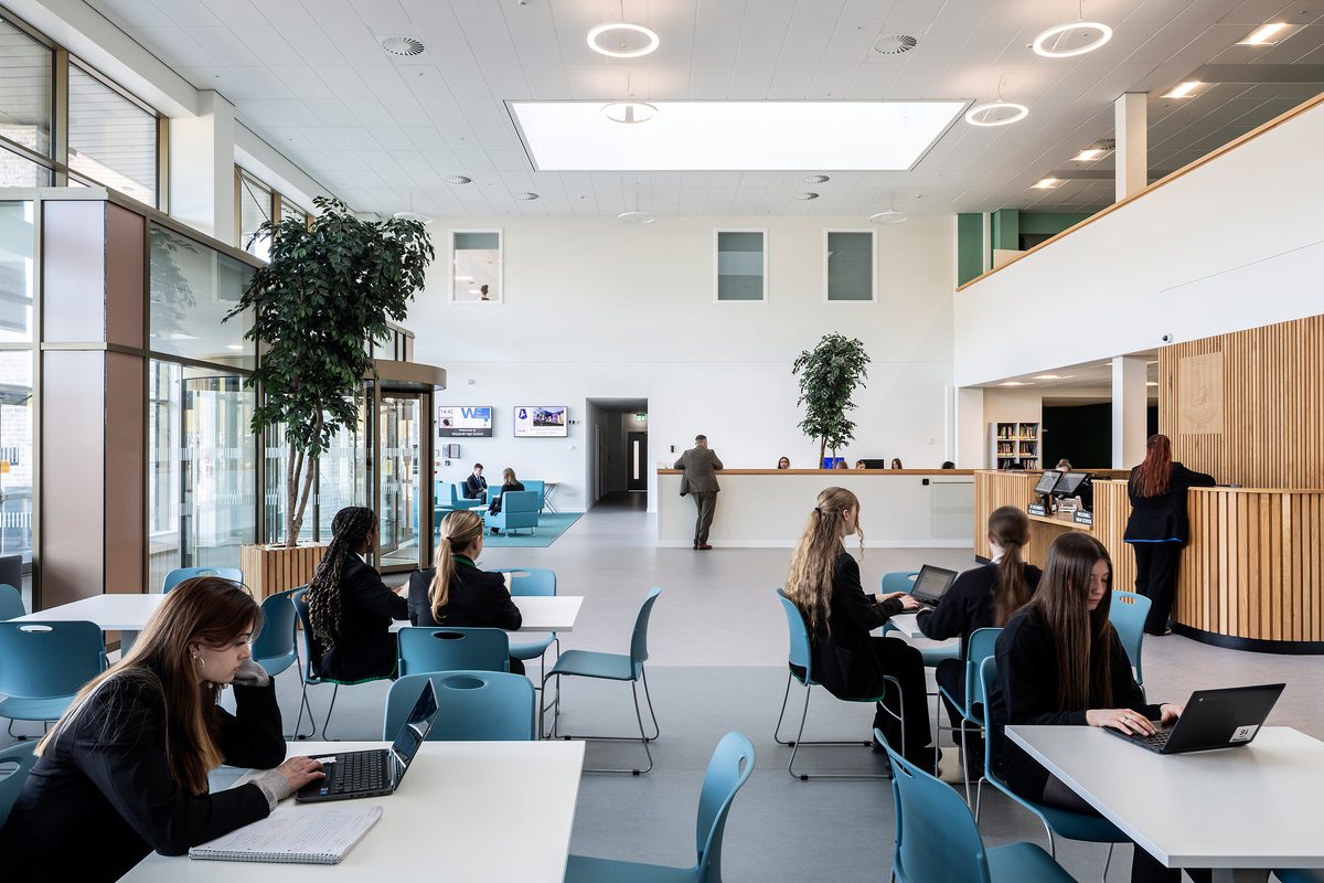 The bright, naturally lit reception area at Woodmill and St Columba's RC High School with pupils sat across various tables working on laptops and a reception desk to the far right.