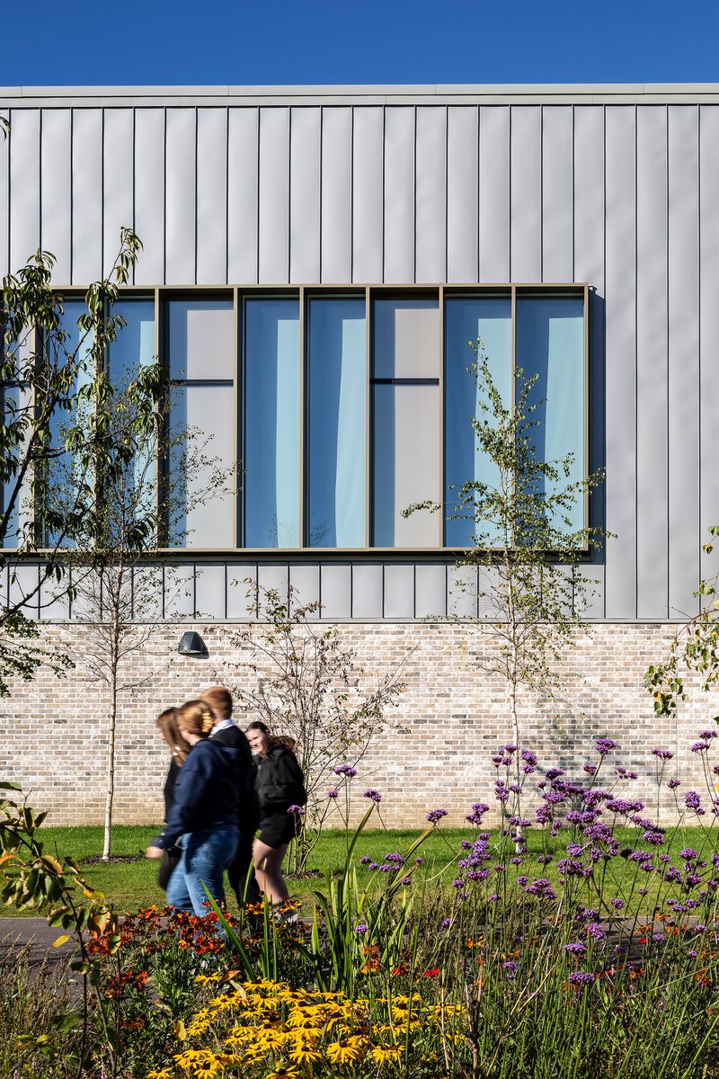 Pupils walking through the communal landscaped exterior of the Woodmill and St Columba's RC High School, with the school in the background and biodiverse planting in the foreground.