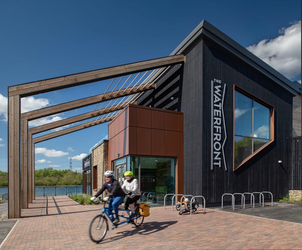 Two people on a tandem bike cycling past the entrance to the Rother Valley Country Park visitor centre on a bright, sunny day.