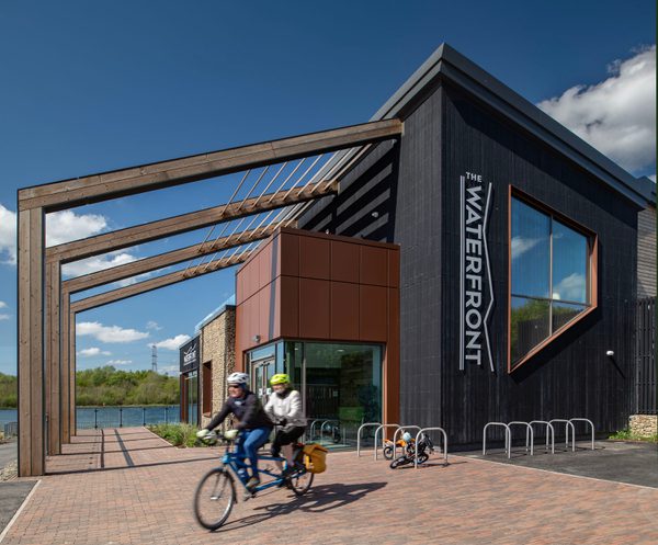 Two people on a tandem bike cycling past the entrance to the Rother Valley Country Park visitor centre on a bright, sunny day.