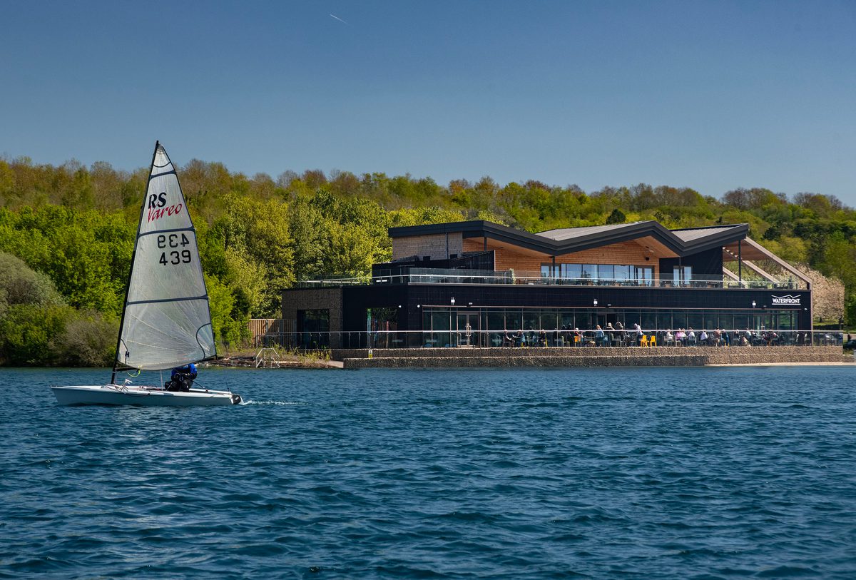 The Waterfront cafe at Rother Valley Country Park situated next to a large lake with a sail boat in the foreground.