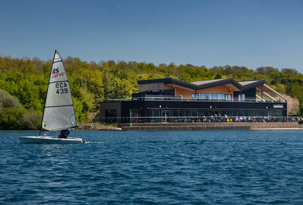 The Waterfront cafe at Rother Valley Country Park situated next to a large lake with a sail boat in the foreground.
