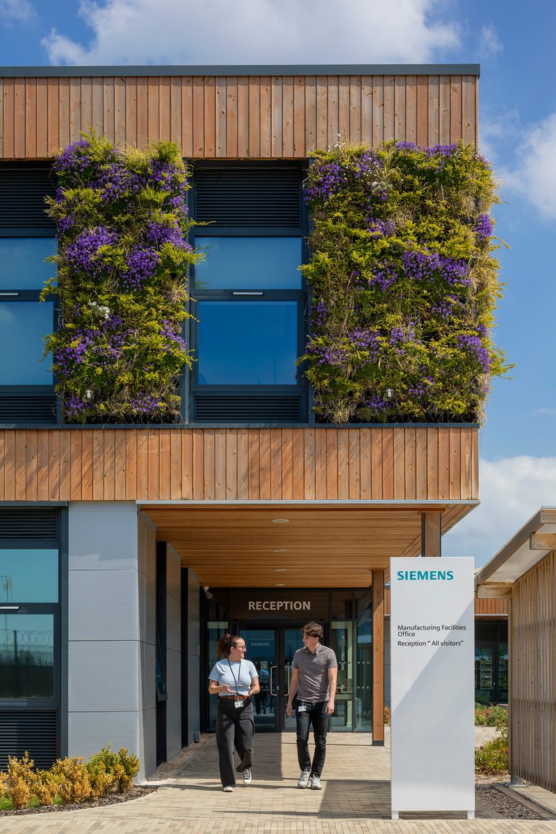 Two people walking in entrance of Siemens train manufacturing facility, with wooden facade and windows surrounded by a green wall.