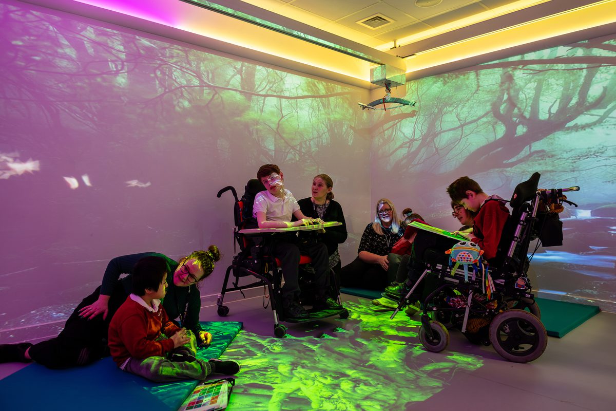 SEND students interacting with teachers in a sensory room with various colours and patterns around them.