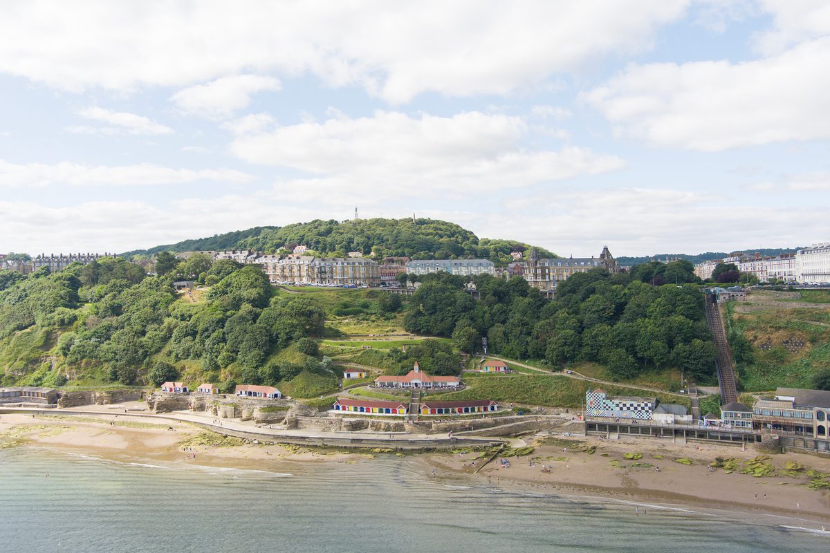 An aerial drone shot of the rebuilt colourful beach chalets on Scarborough's South Cliff taken from afar, showing their cliffside, beach front location.