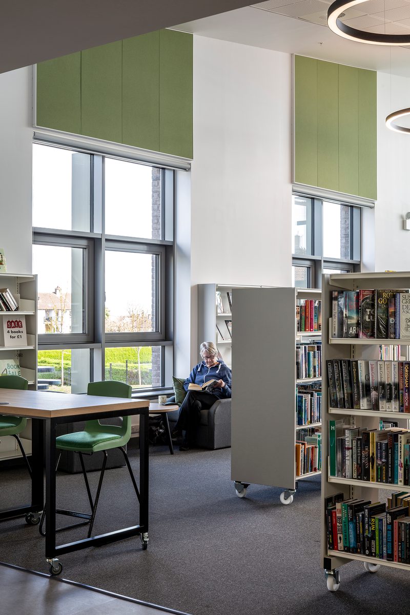A portrait photograph of the community library filled with shelves filled with books and a person sat down reading against a large window.