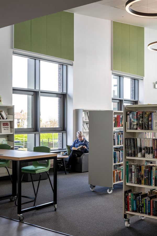 A portrait photograph of the community library filled with shelves filled with books and a person sat down reading against a large window.