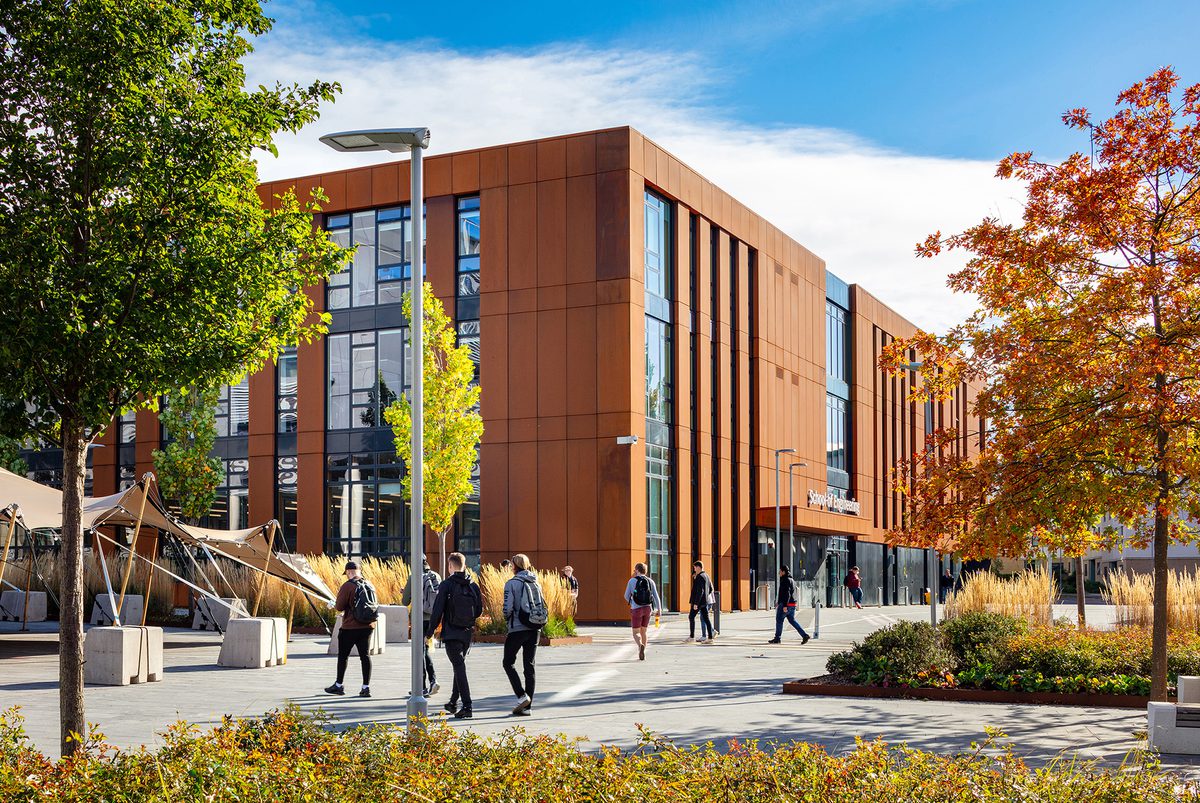 Rust coloured exterior facade of the School of Engineering, with triple height glazing, enhanced public realm and students walking around the building.